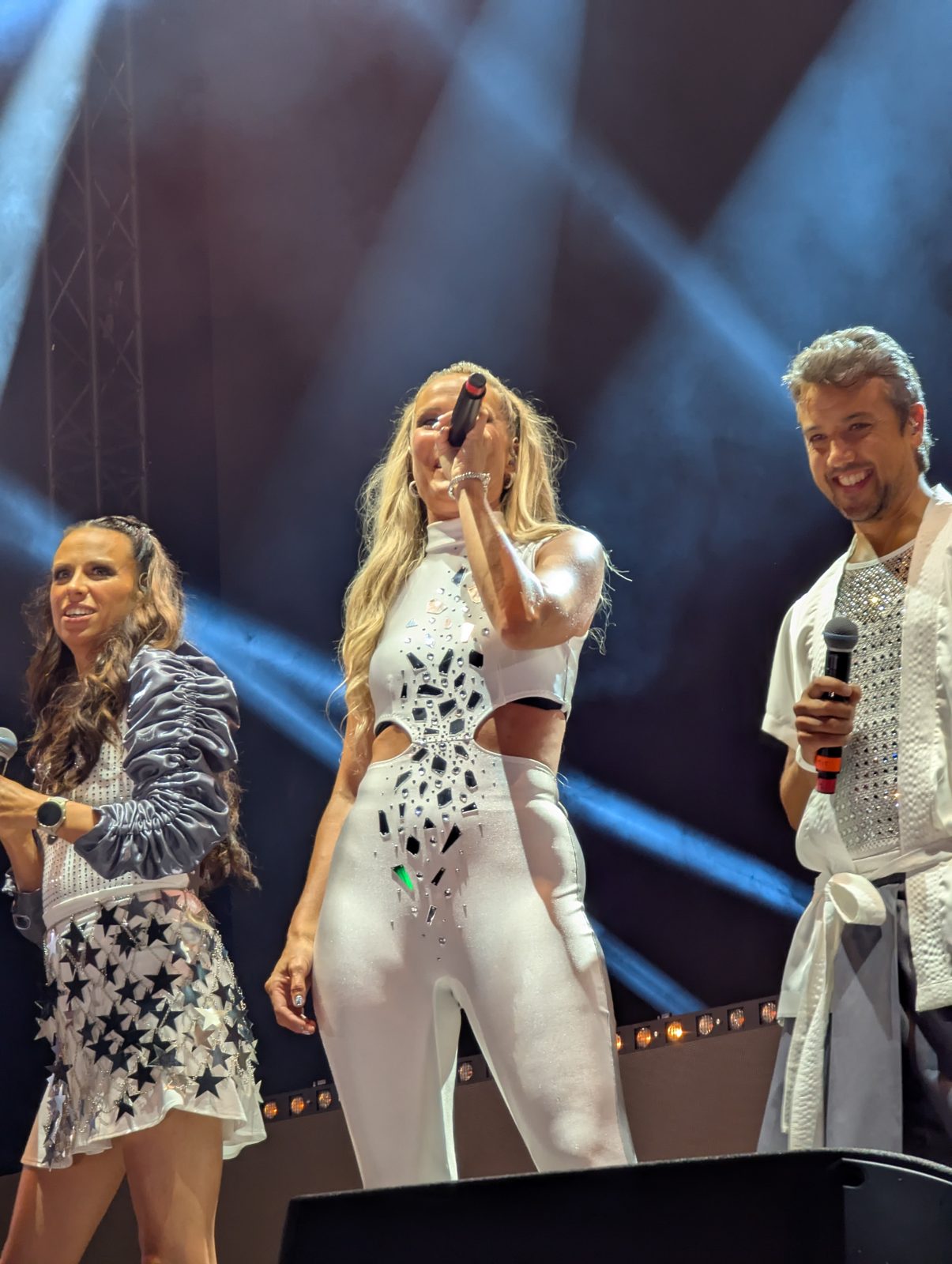 Three A*Teens members smiling together under blue stage lighting in Stockholm, 2025
